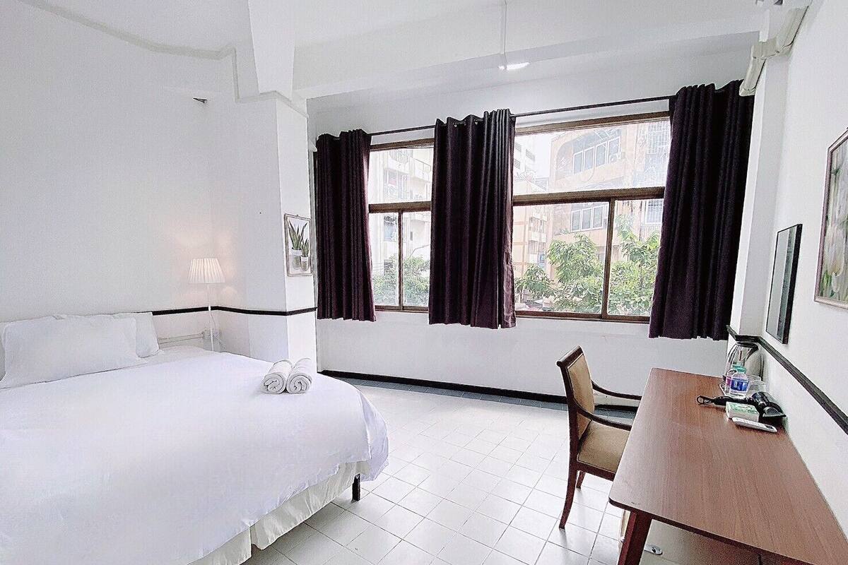 Ein Hotelzimmer mit einem Bett, einem Schreibtisch und einem Stuhl. Es gibt Fenster mit Vorhängen, eine Lampe und eine Pflanze an der Wand.