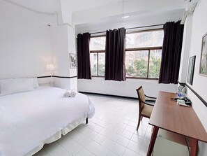 Hotelkamer met een bed, een bureau en een stoel. Er zijn ramen met gordijnen, een lamp en een plant aan de muur.