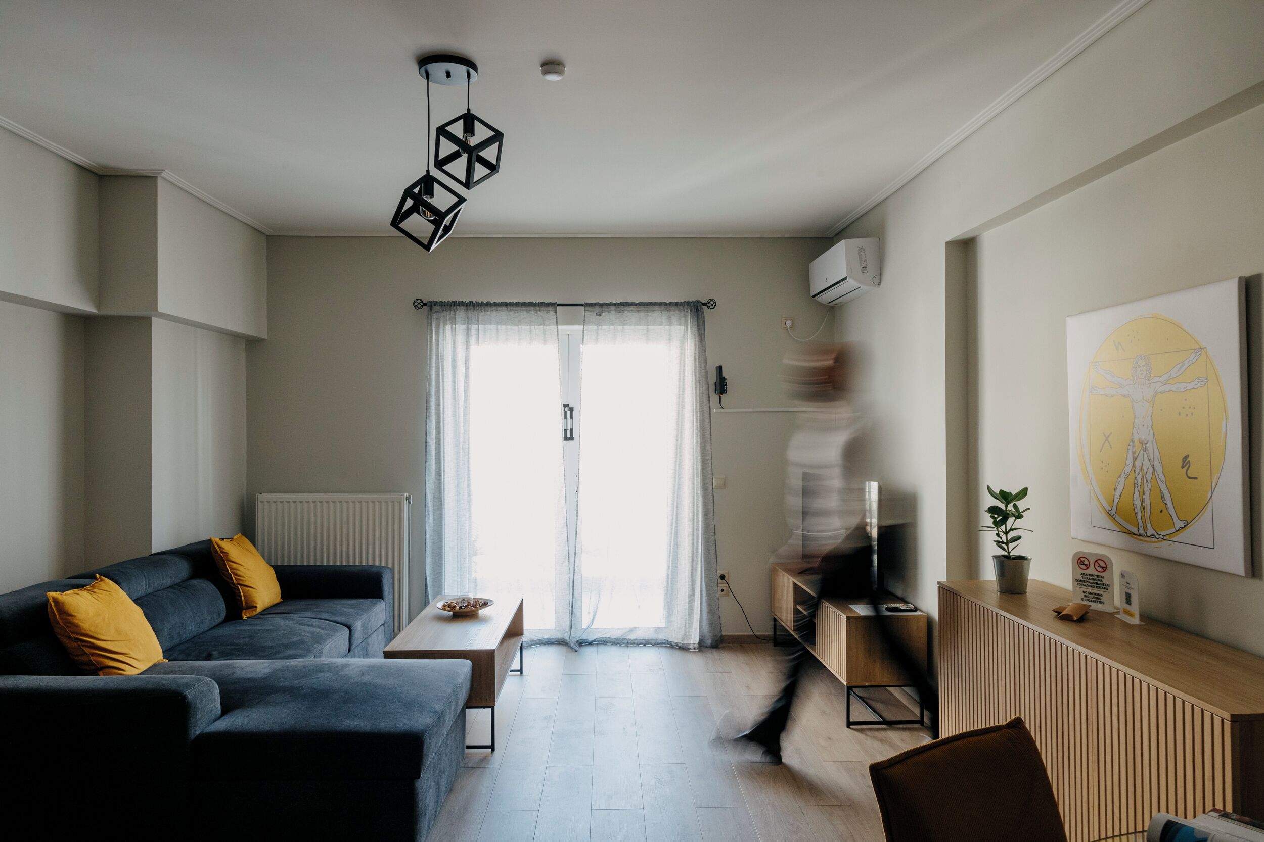 Un salon moderne avec un canapé, une table basse et un téléviseur. Il y a une fenêtre avec des rideaux, une plante sur une table d’appoint et un tableau au mur.