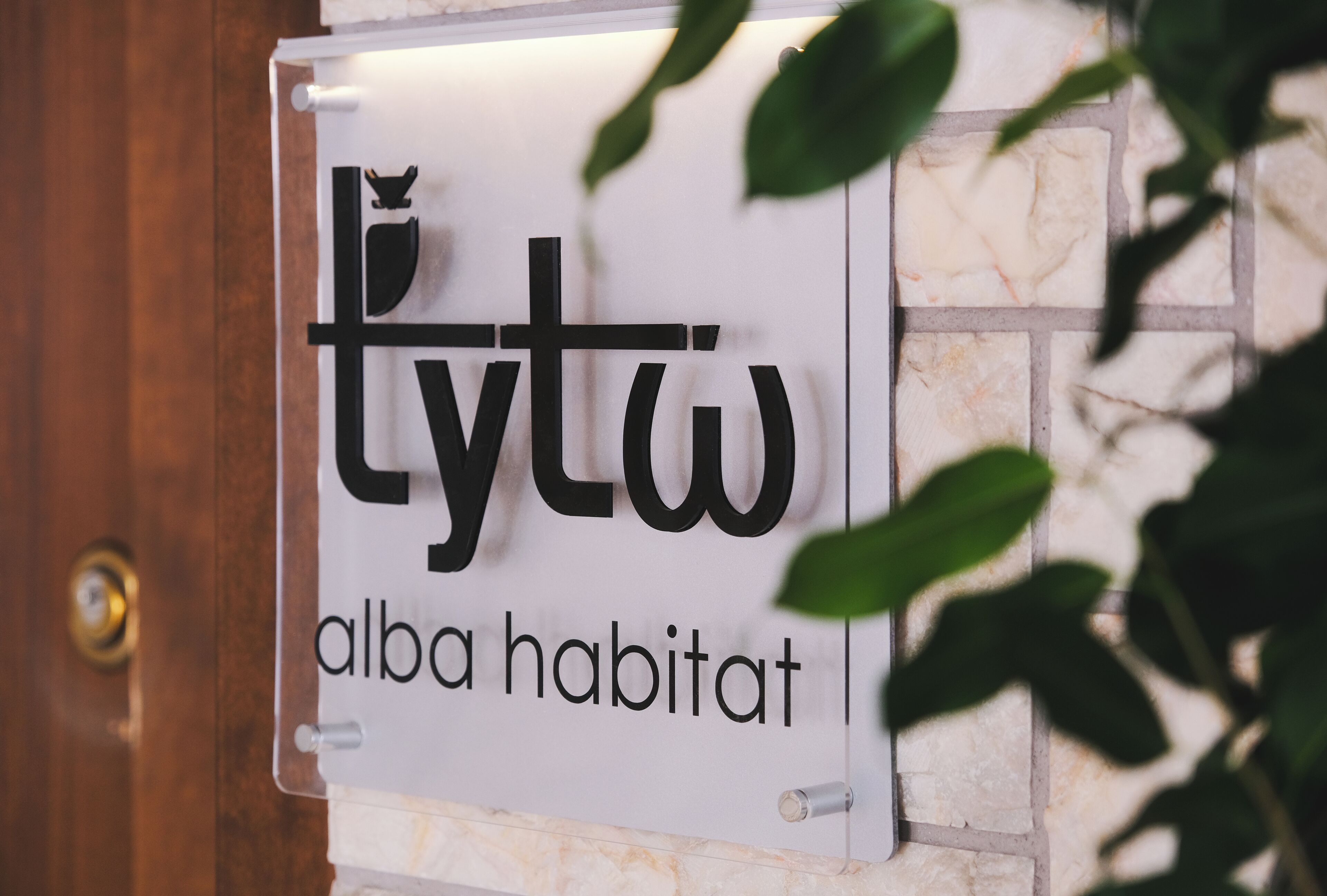 Une enseigne indiquant « tytwa alba habitat » est fixée à un mur, à côté d’une porte en bois et de carreaux de marbre.