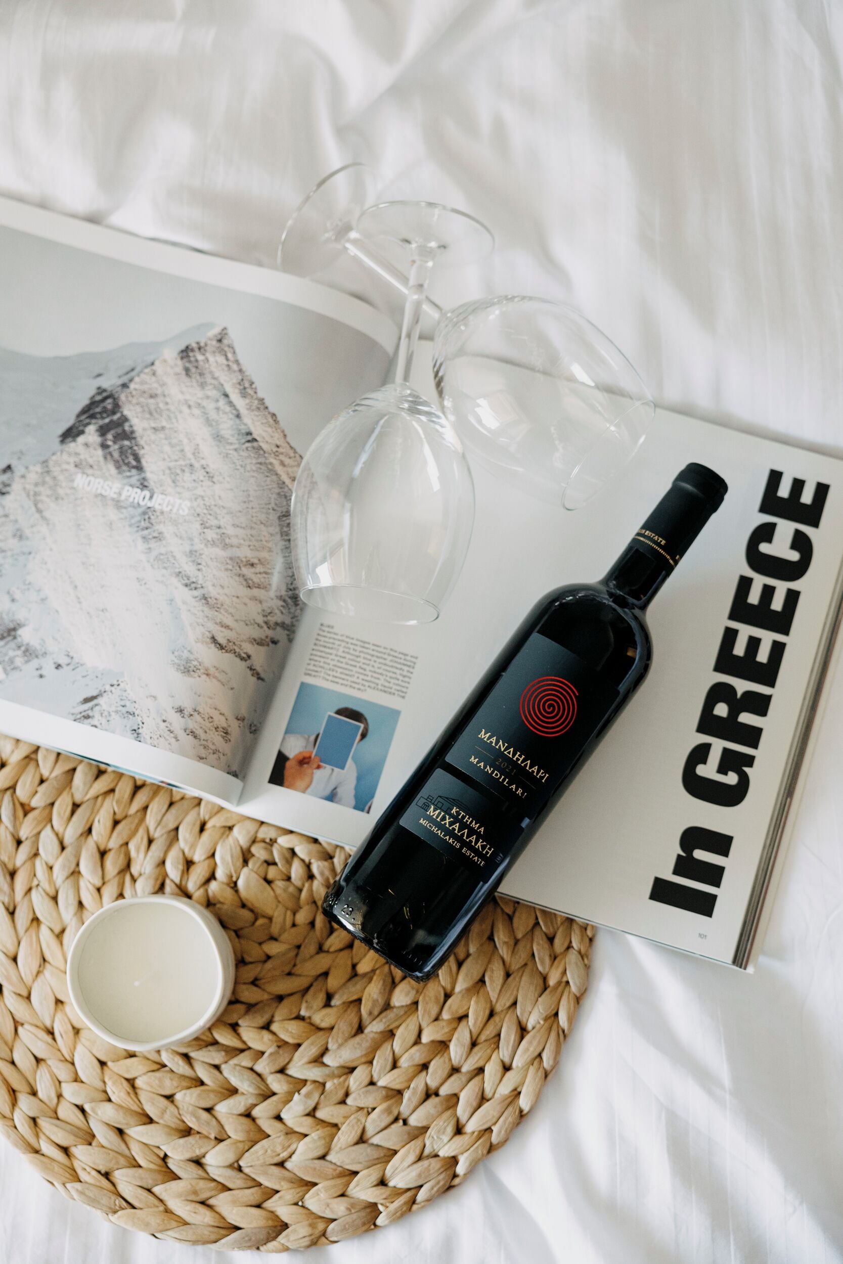Una bottiglia di vino Manosha, un sottotavolo intrecciato, un bicchiere e una rivista intitolata 'In Greece'.