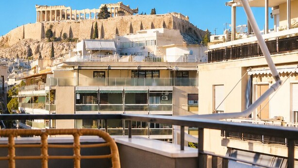 Un balcon agrémenté de meubles en osier, avec vue sur des immeubles modernes et, au loin, sur des ruines antiques.