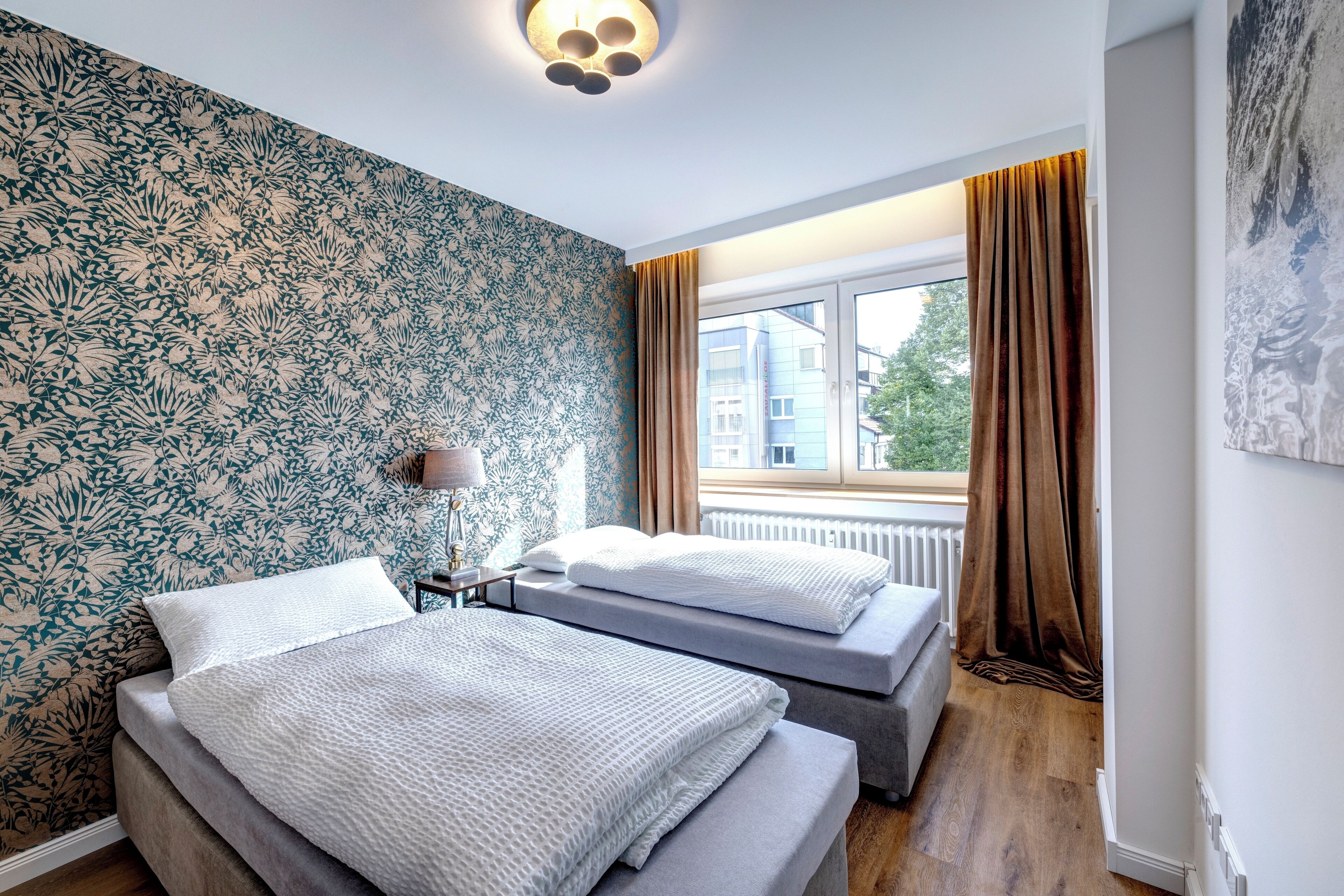 Une chambre avec deux lits, une fenêtre avec des rideaux, un radiateur et un mur orné d’un motif floral.
