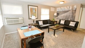 A living room with a sofa, a coffee table, a dining table with chairs, a lamp, and a painting on the wall. - 1 Bedroom Apartment Near Notre Dame 1309 5 (South Bend)