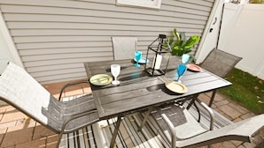 Um pátio externo com uma mesa e cadeiras de metal escuro, um candeeiro e um vaso com planta.