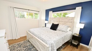Quarto com cama, mesa de cabeceira, abajur, janela com cortinas e tapete estampado.