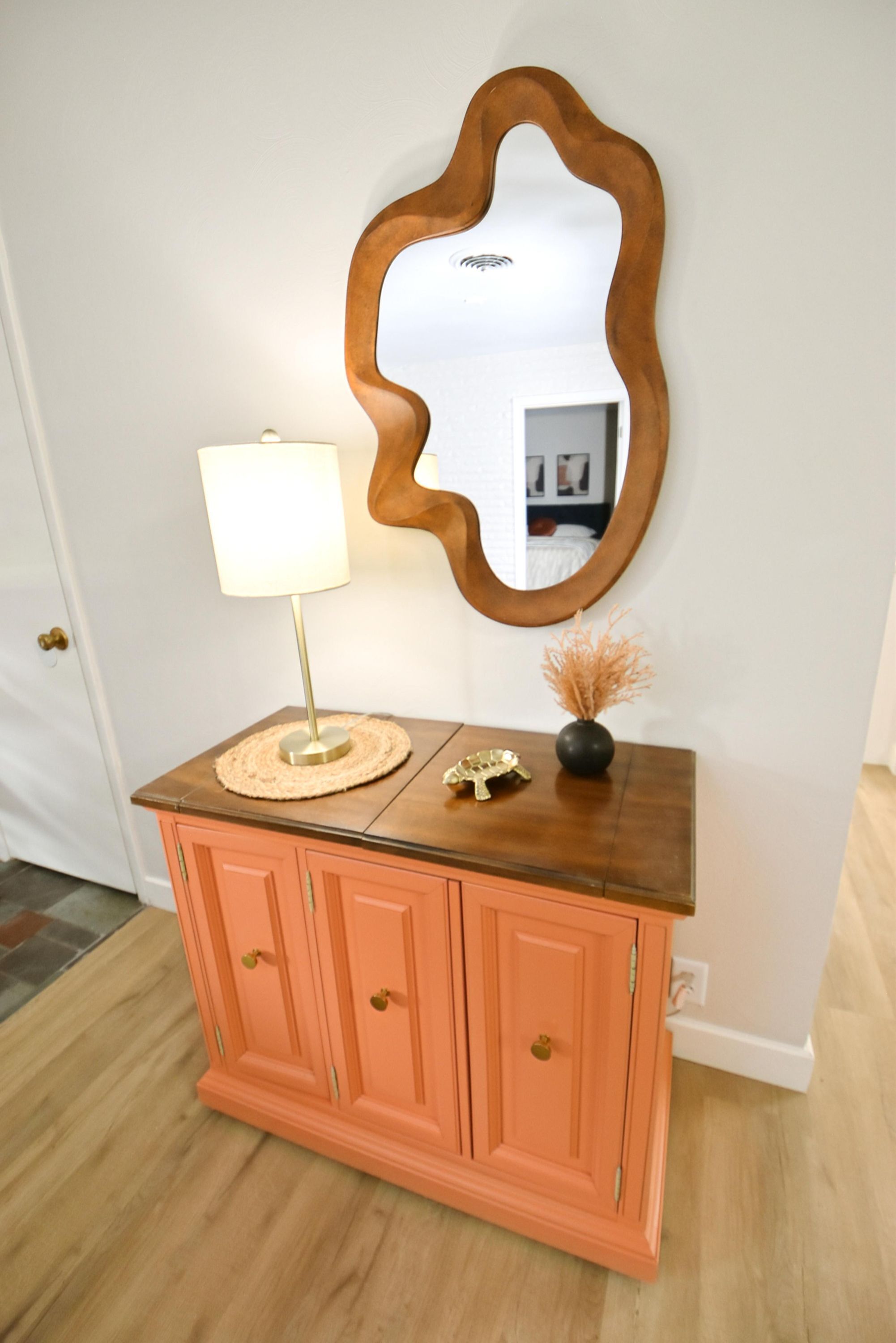A pink cabinet with a lamp and decorative items on top, a mirror with a wavy frame, and a door in the background.
