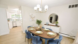 Un'area pranzo con un tavolo di legno apparecchiato per sei persone, sedie blu, un lampadario, uno specchio rotondo e una cucina sullo sfondo.