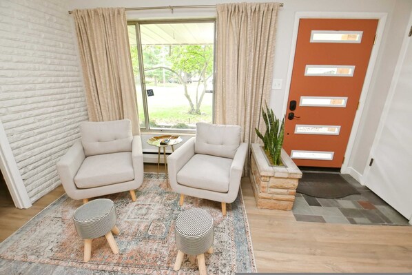 Un salon moderne avec deux canapés, une table basse et deux poufs. On y trouve une grande fenêtre avec des rideaux, une porte avec un judas et une plante en pot.