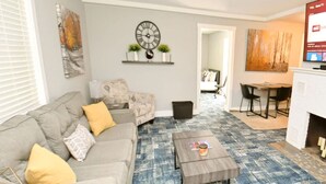 Ein modernes Wohnzimmer mit einem grauen Sofa, einem Couchtisch und einem blauen Teppich mit Muster. An der Wand hängt eine Uhr und über dem Sofa ein Bild.