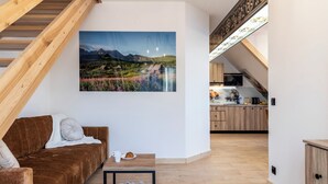 Un loft moderno con una escalera de madera, una zona de estar acogedora con un sofá marrón, un cuadro de un paisaje montañoso, y una cocina al fondo.