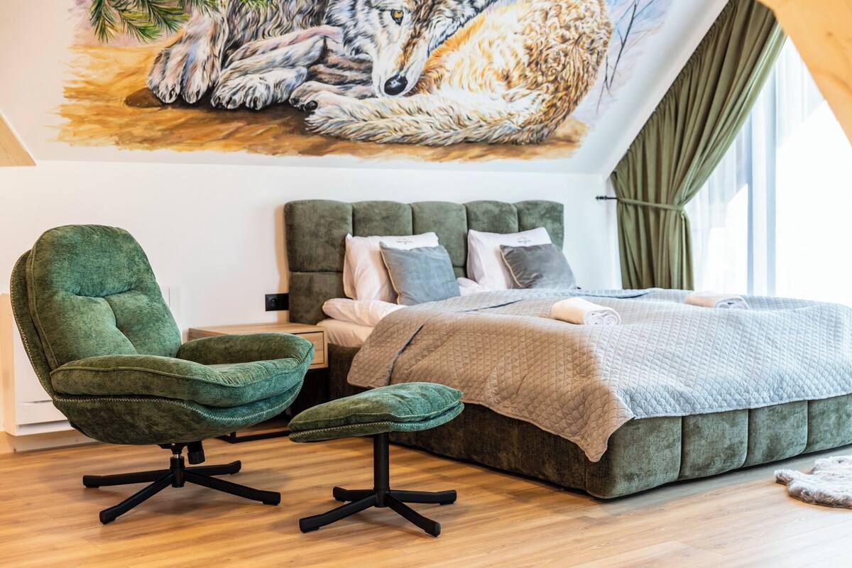 A bedroom with a green upholstered chair, a bed with a quilted cover, and a mural of a wolf on the wall.