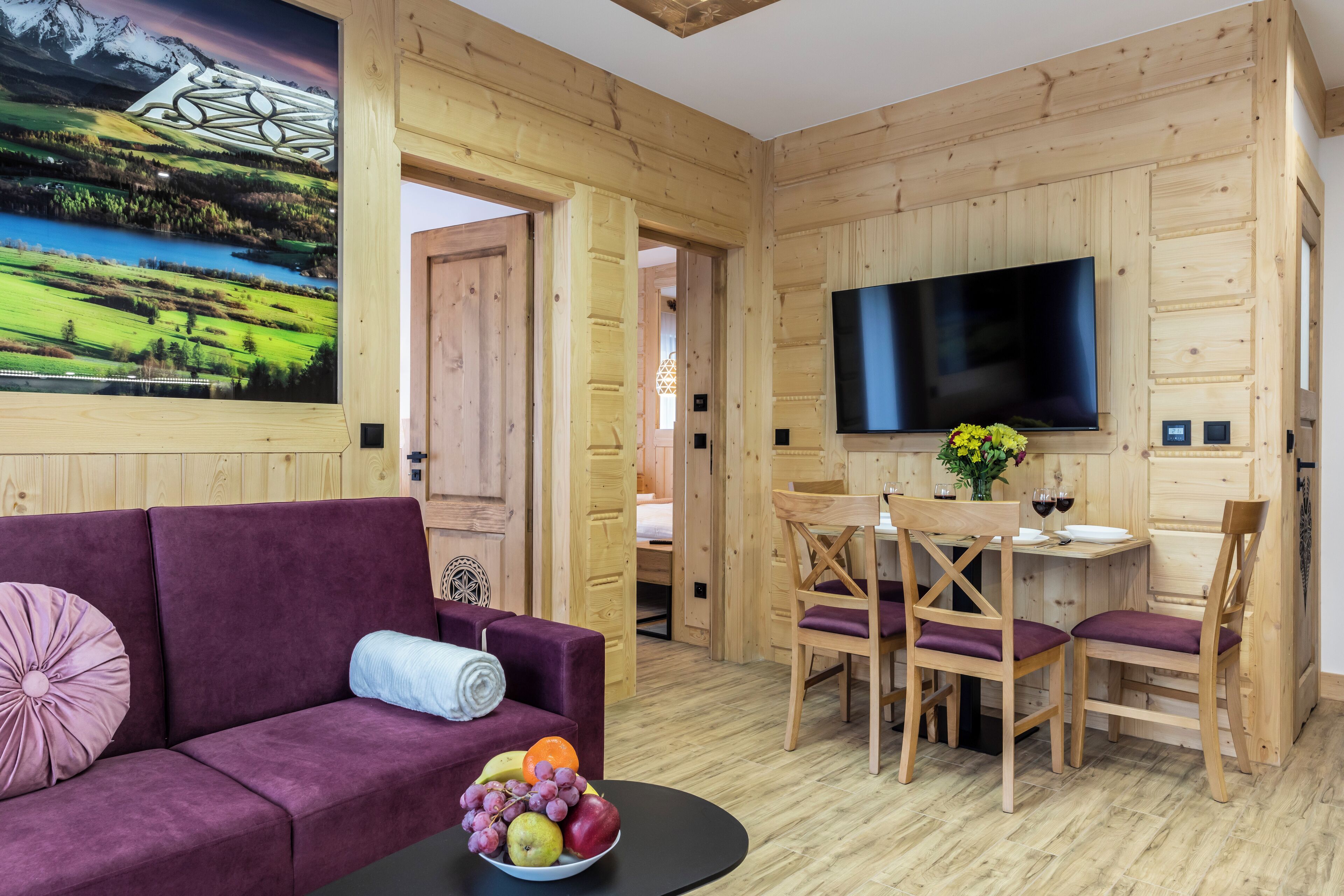 Un intérieur en bois avec un canapé violet, une table à manger et des chaises. Un téléviseur à écran plat est fixé au mur.