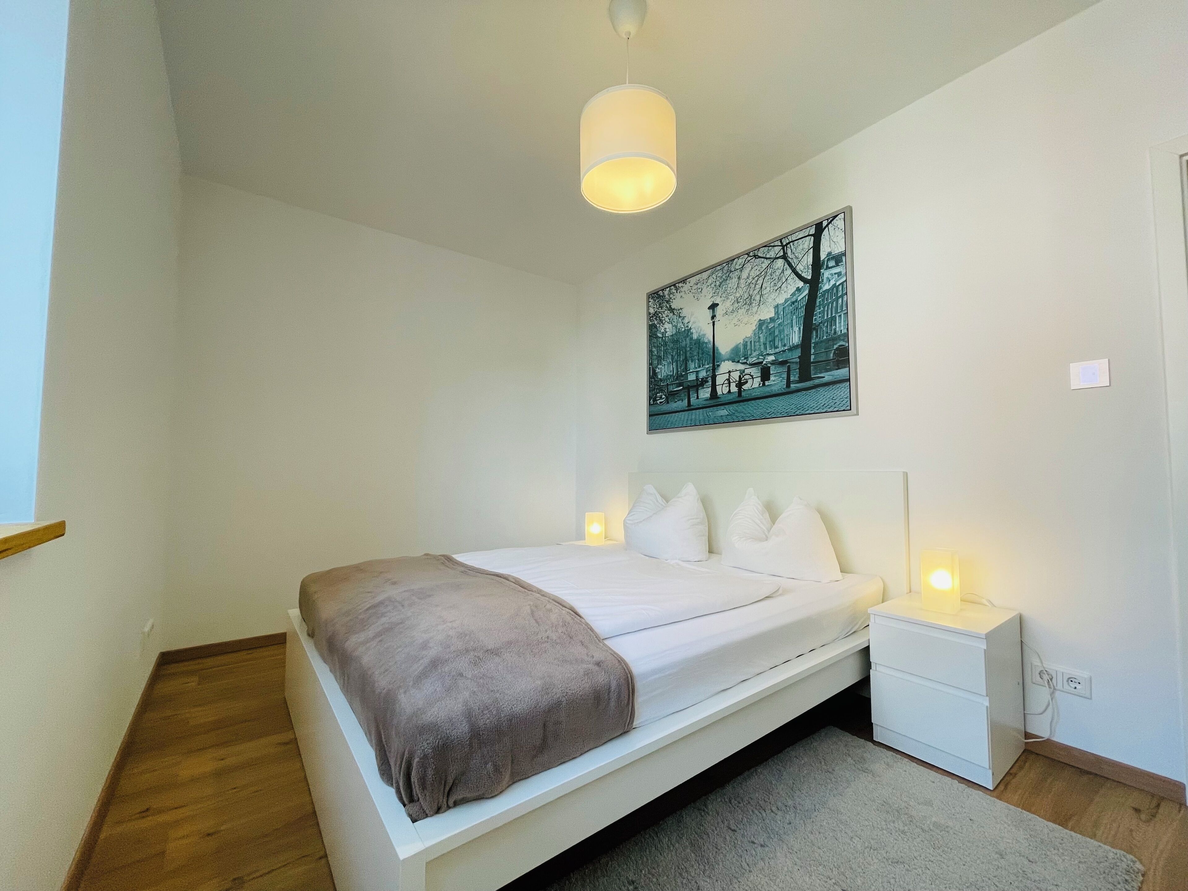 A bedroom with a bed, bedside tables, a nightstand, a painting, and a hanging light fixture.