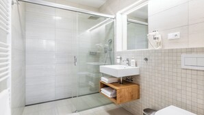 Un baño moderno con ducha envuelta en vidrio, lavamanos blanco y tocador de madera.
