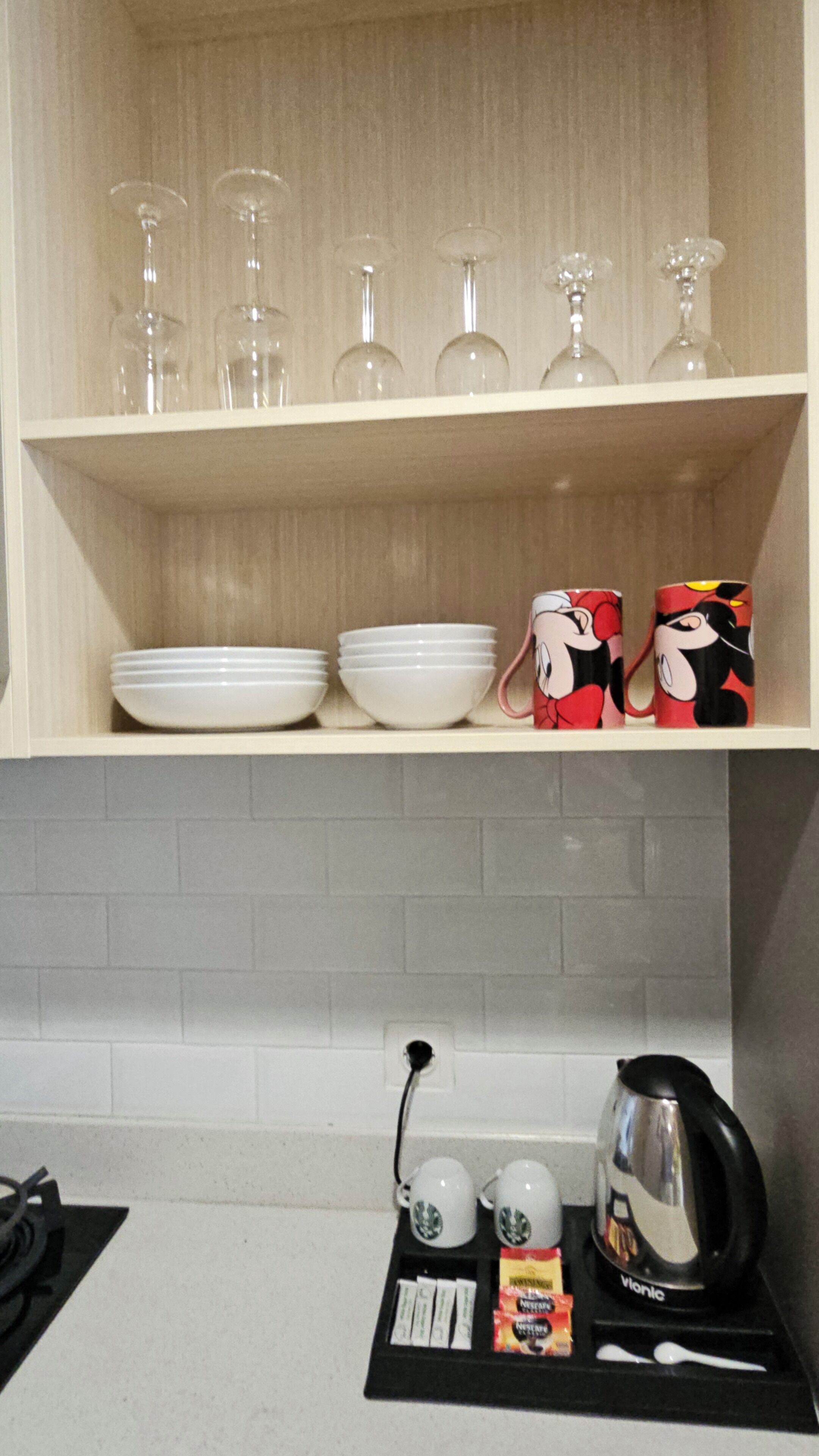 A kitchen with a shelf holding wine glasses, bowls, and mugs. Below, there is a coffee maker, cups, and condiments.