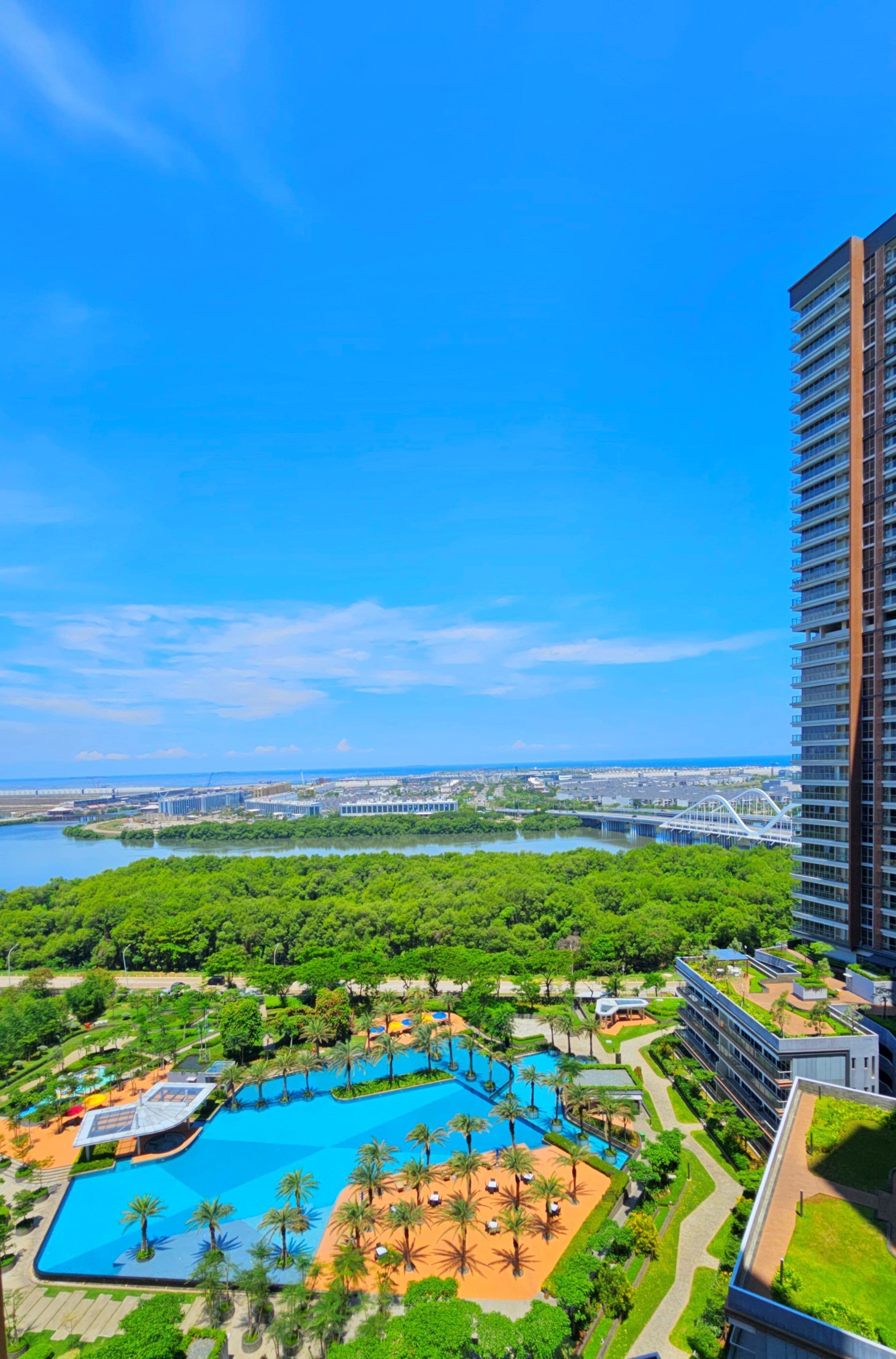 A resort with a large swimming pool surrounded by palm trees and greenery, a bridge in the background, and a tall building to the right.