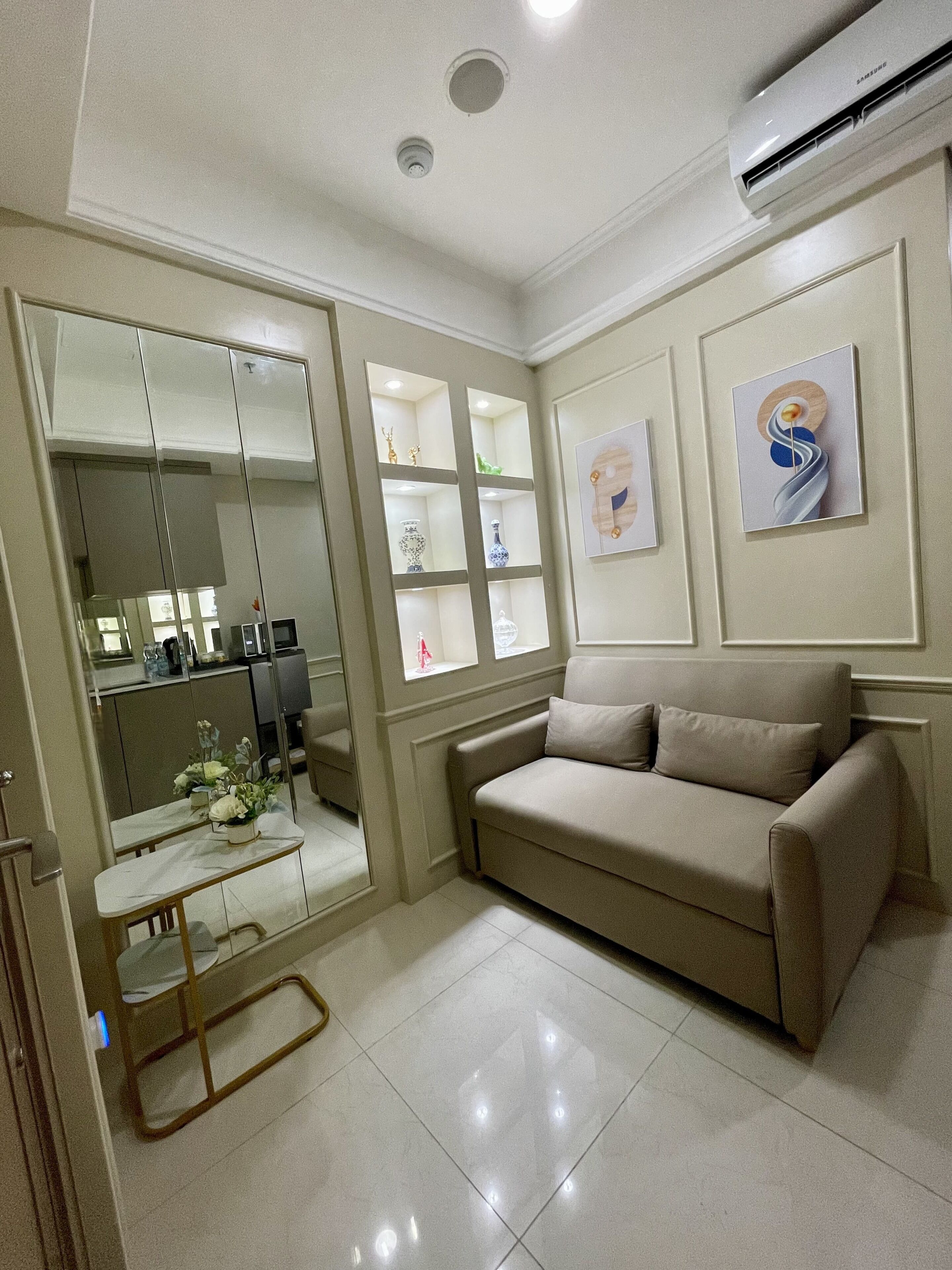 A modern living room with a beige sofa, a glass coffee table, and a mirrored wardrobe. There is a built-in shelf unit with decorative items and a wall-mounted air conditioner.