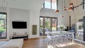 A modern living room with a large flat-screen TV, a sofa, a dining table with clear chairs, and a large window with a view of the outdoors. - Waterlane Riverside - SPA, Pool, Gym & Parking by Downtown Apartments (Gdansk)