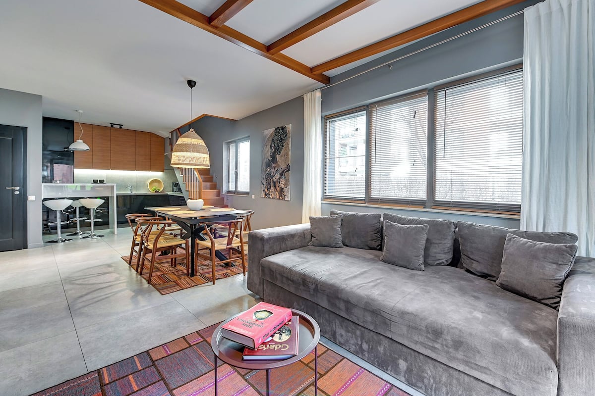 A modern living room with a grey sofa, a dining table with chairs, and a kitchen in the background.