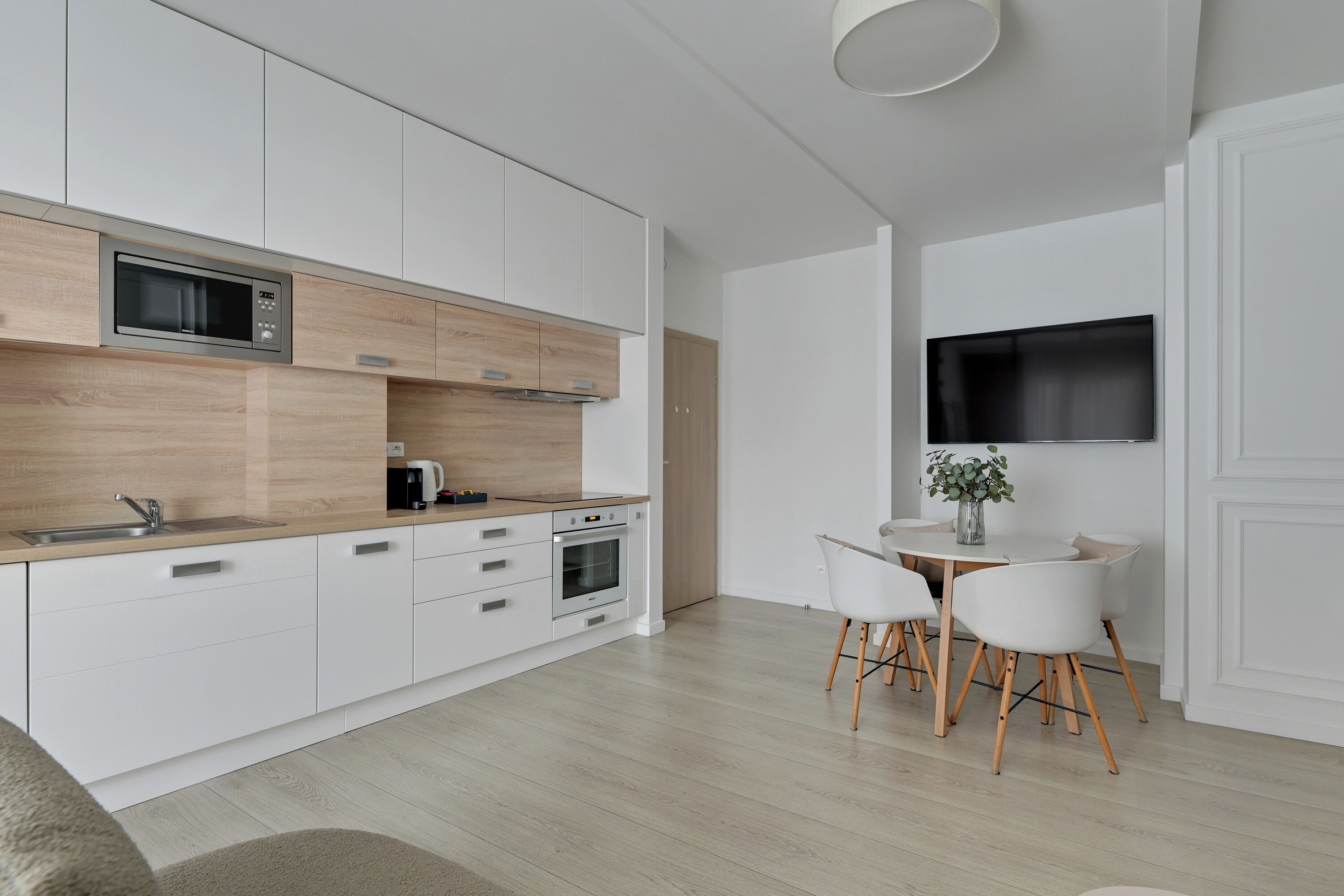 A modern kitchen with white cabinetry, a built-in microwave, and an oven. There is a dining area with a table and chairs.