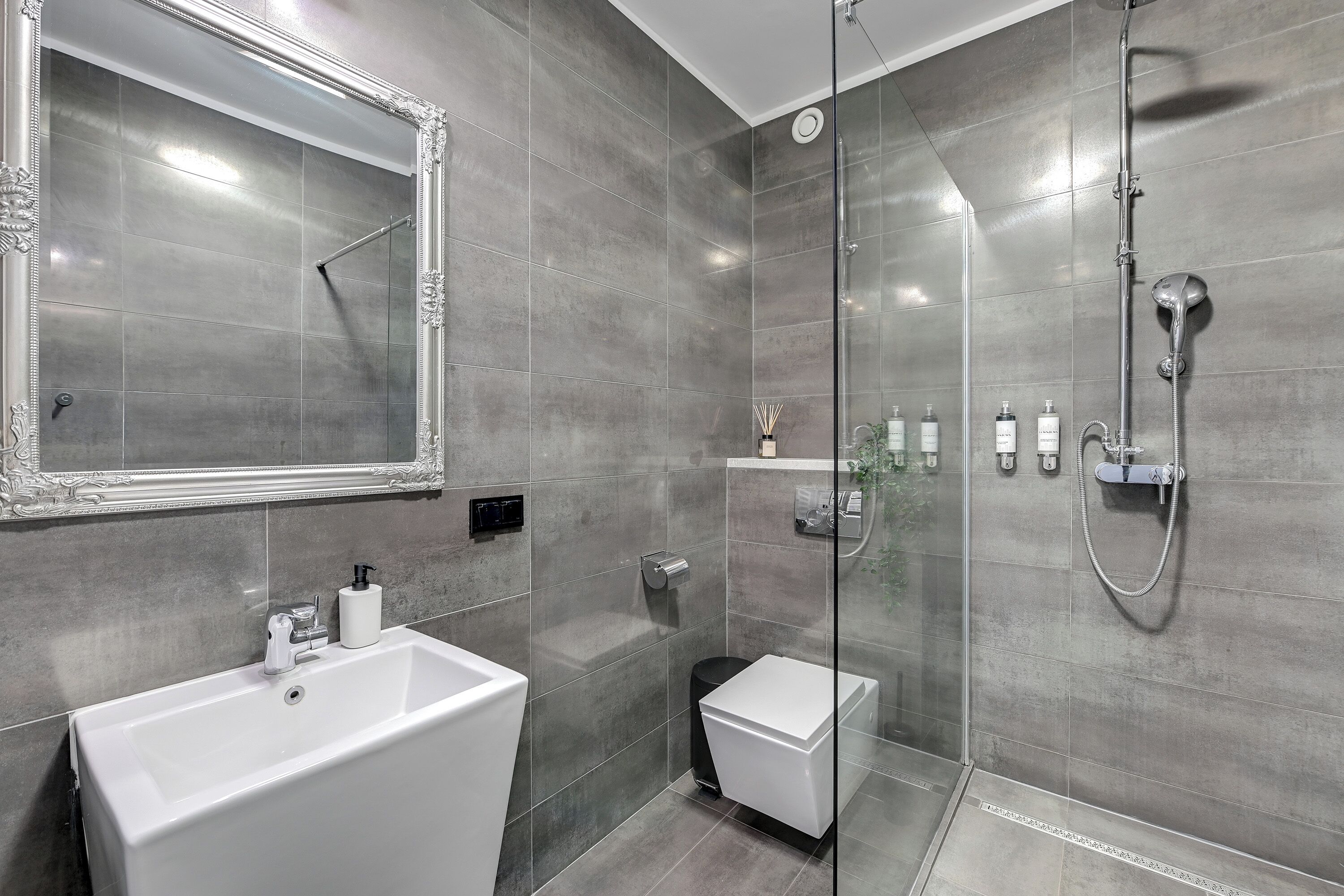 Une salle de bain moderne avec un grand miroir, un lavabo blanc, une douche à l’italienne et de petites toilettes.