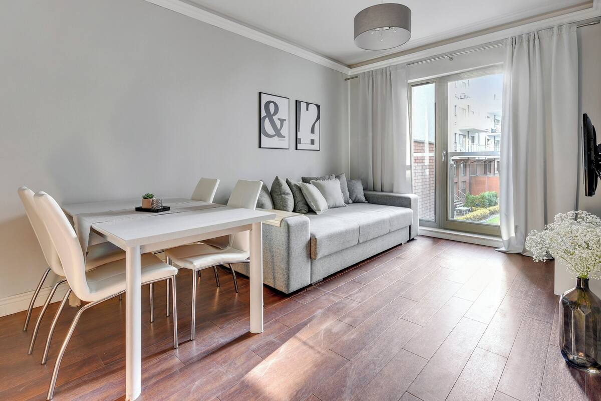 A modern living room with a grey sofa, white dining table, and chairs. There are two framed pictures on the wall, a large window with curtains, and a vase with flowers on the floor.
