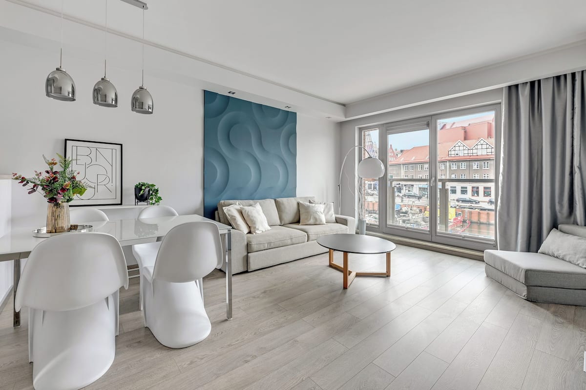A modern living room with a sofa, dining table, and chairs, large windows with a city view, and pendant lights.