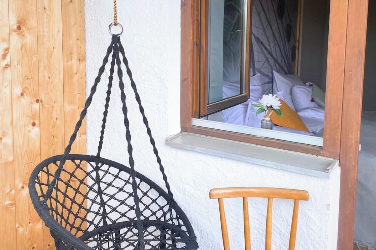 Ett rum med en hängstol, en trästol och ett fönster med utsikt över en säng och en vas med blommor.