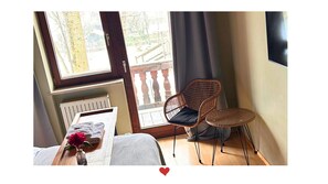 A room with a bed, a chair, a small table with a vase, and a view of a balcony.