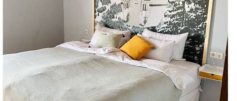 Una cama bien hecha con sábanas blancas y una sola almohada amarilla, una mesita de noche con toma de corriente, y un cuadro colgado en la pared que representa un paisaje montañoso.