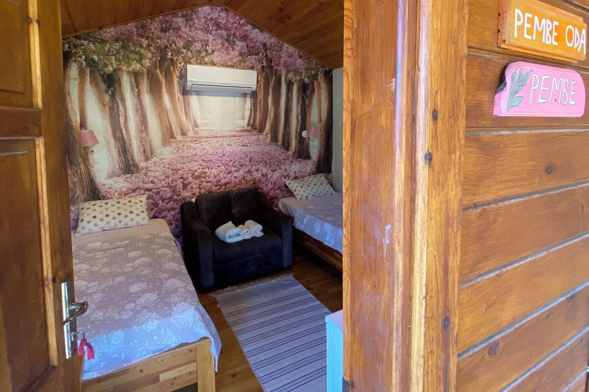 Ein gemütliches Blockhauskabinenzimmer mit Holzwänden, einem Bett, einem Sessel und einem kleinen Tisch. An der Wand befinden sich Schilder mit dem Zimmernamen.