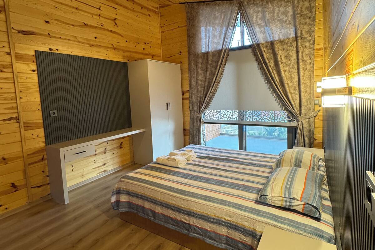 Ein Schlafzimmer einer Holzhütte mit einem Bett, einem Schminktisch, einem an der Wand montierten Fernseher und einem Fenster mit Vorhängen.