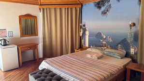 Un dormitorio con cama, escritorio, silla, refrigerador pequeño y vista al mar a través de un ventanal grande.