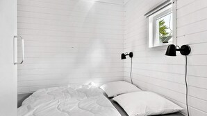 Een enkel bed met een witte matras en kussens, een klein raam met uitzicht op groen, en twee zwarte, aan de muur bevestigde lampen.
