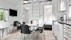 Een moderne keuken met witte kasten, een eethoek met zwarte stoelen, een aan de muur bevestigde televisie en een raam met uitzicht op groen buiten.