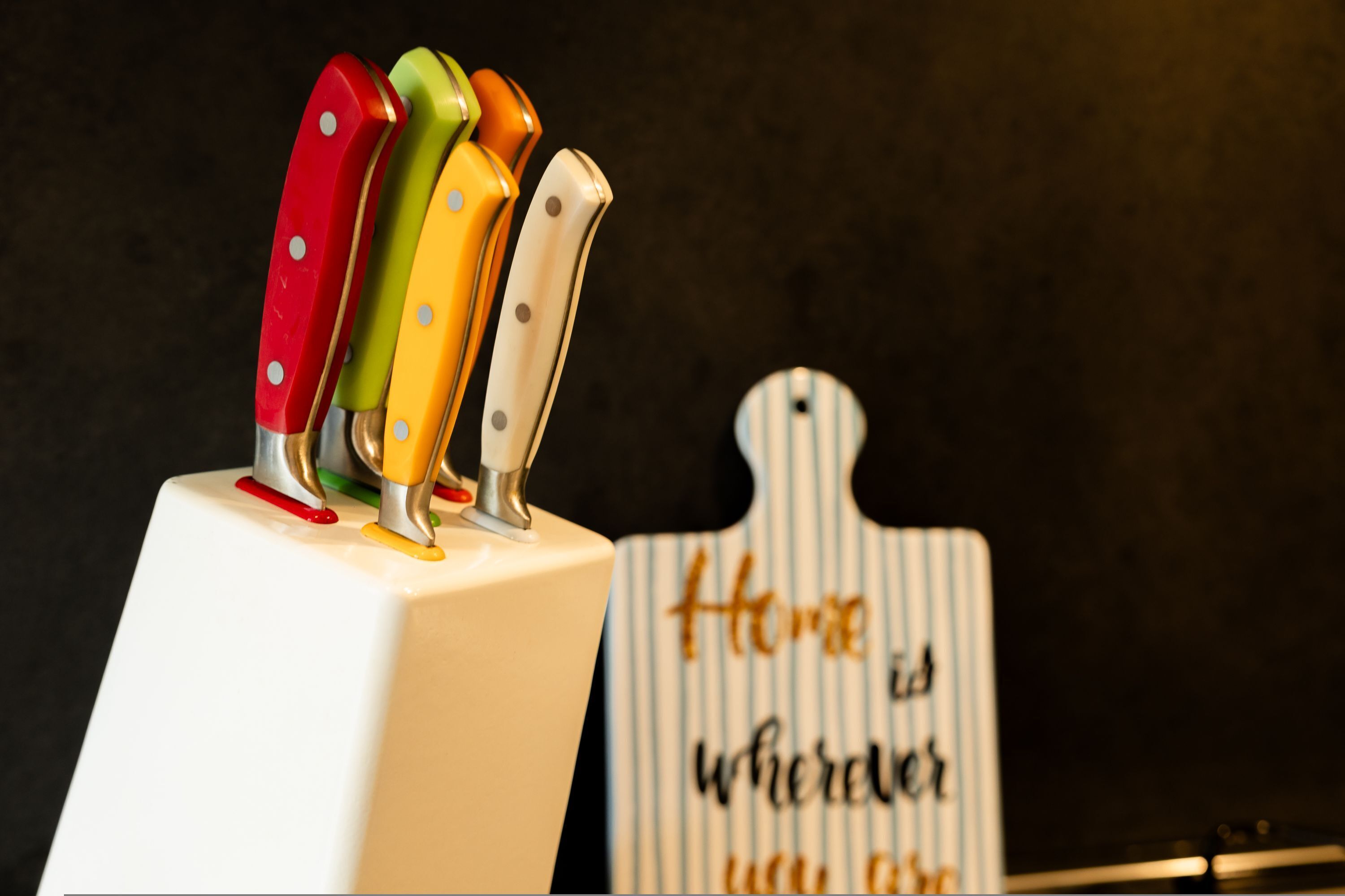 A set of colorful knives on a white stand with a sign in the background that reads "Home is where you are."