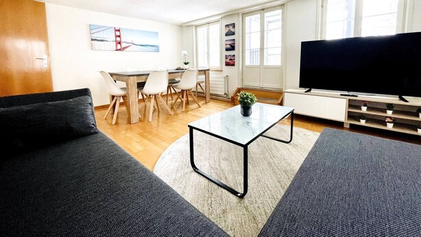 A modern living room with a sofa, a coffee table, a TV, and a dining area with chairs and a table. - Beautiful Old Building Apartment In The Center (St. Gallen)