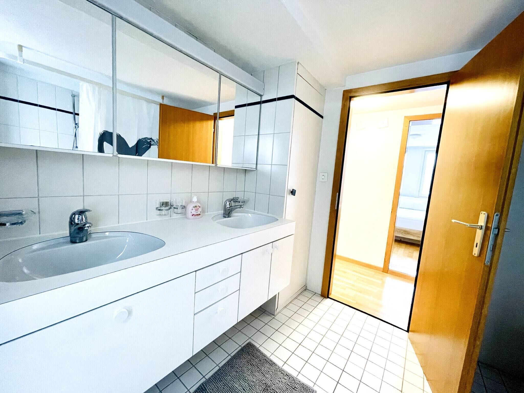 A modern bathroom with two sinks, a large mirror, and a wooden door leading to another room.