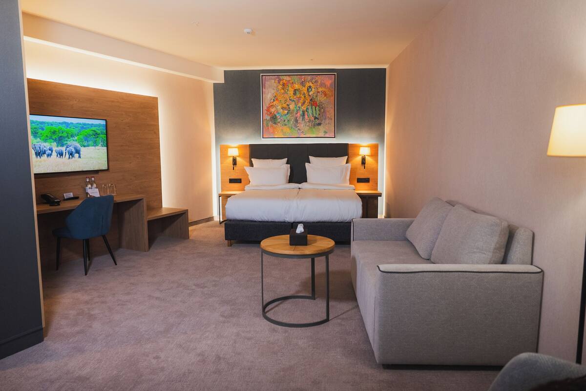 A hotel room with a bed, a desk, a sofa, a TV, and a painting on the wall.