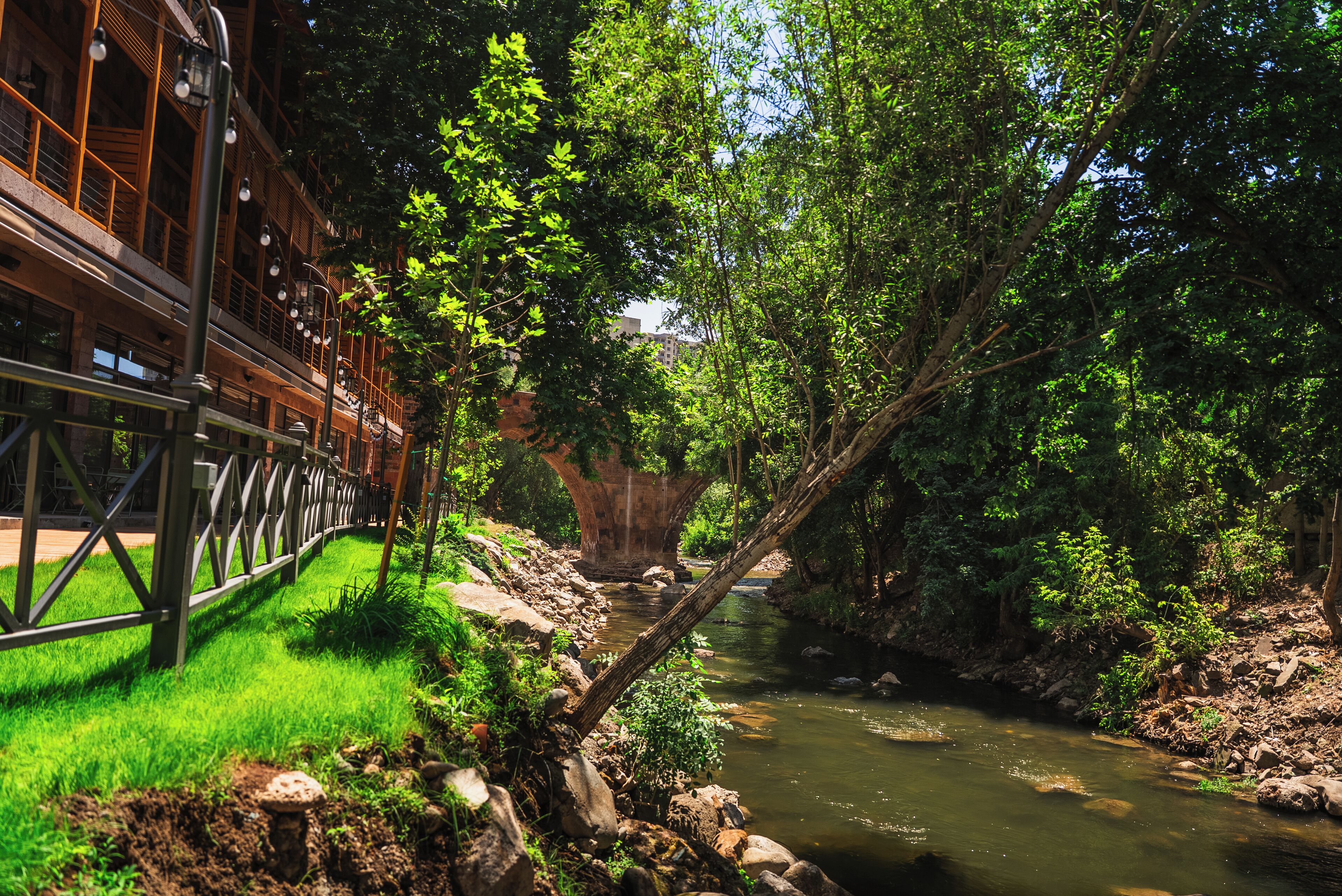 A riverside walkway with a wooden bridge, lush greenery, and a rocky riverbank.