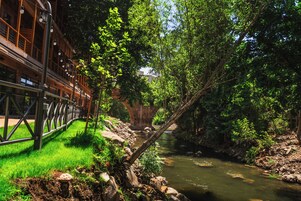 Een promenade langs de rivier met een houten brug, weelderige begroeiing en een rotsachtige oever.