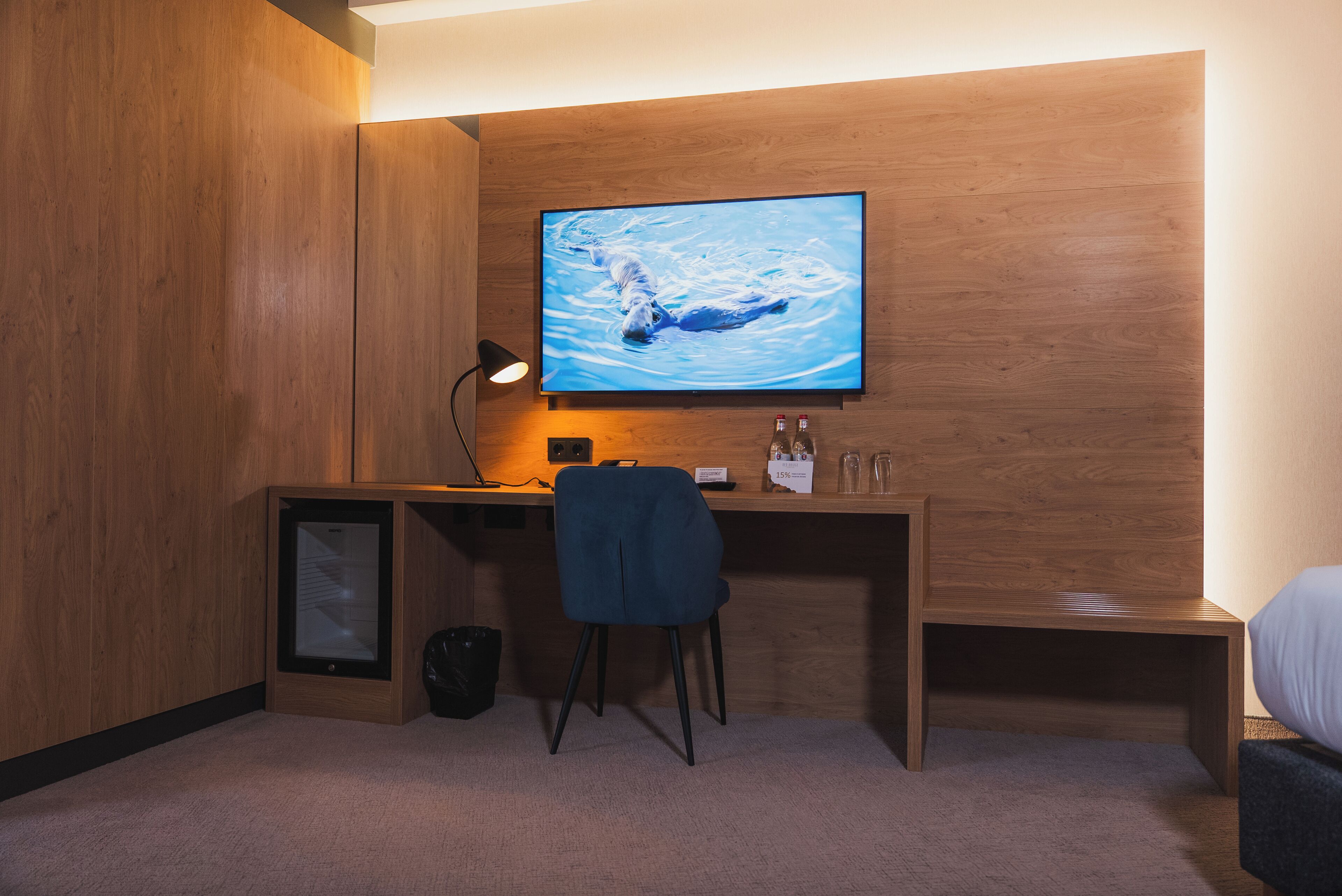 A modern room with a wooden desk, a chair, a television mounted on the wall, and a small shelf with bottles and glasses.