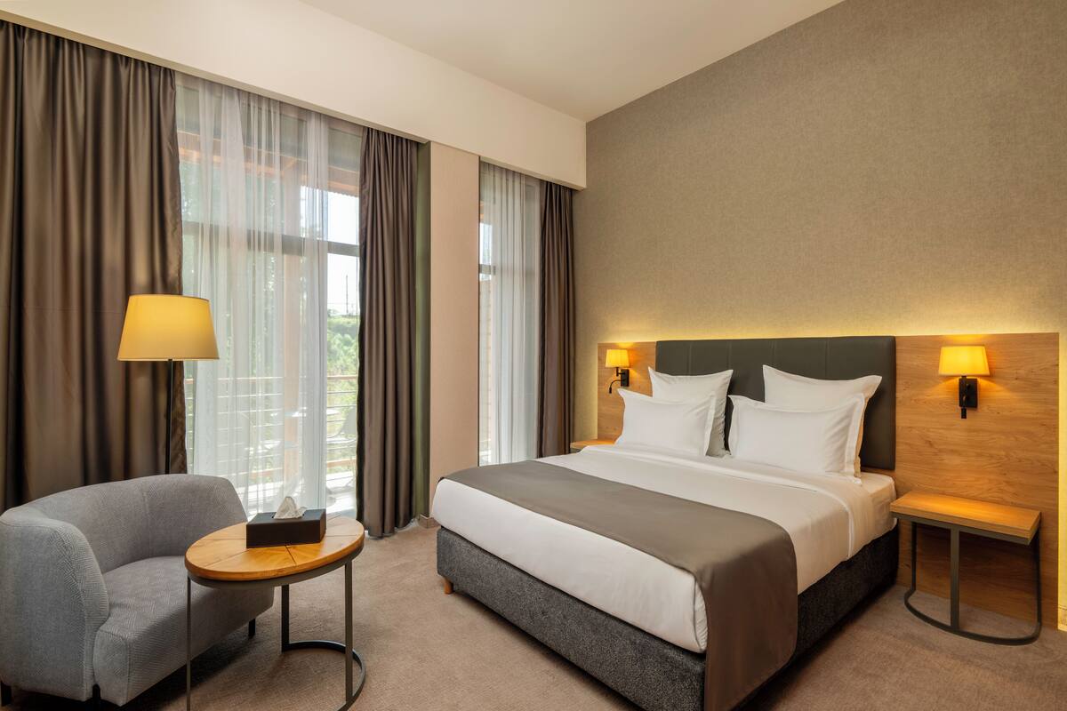 A hotel room with a bed, a chair, a small table, and a nightstand with a lamp.