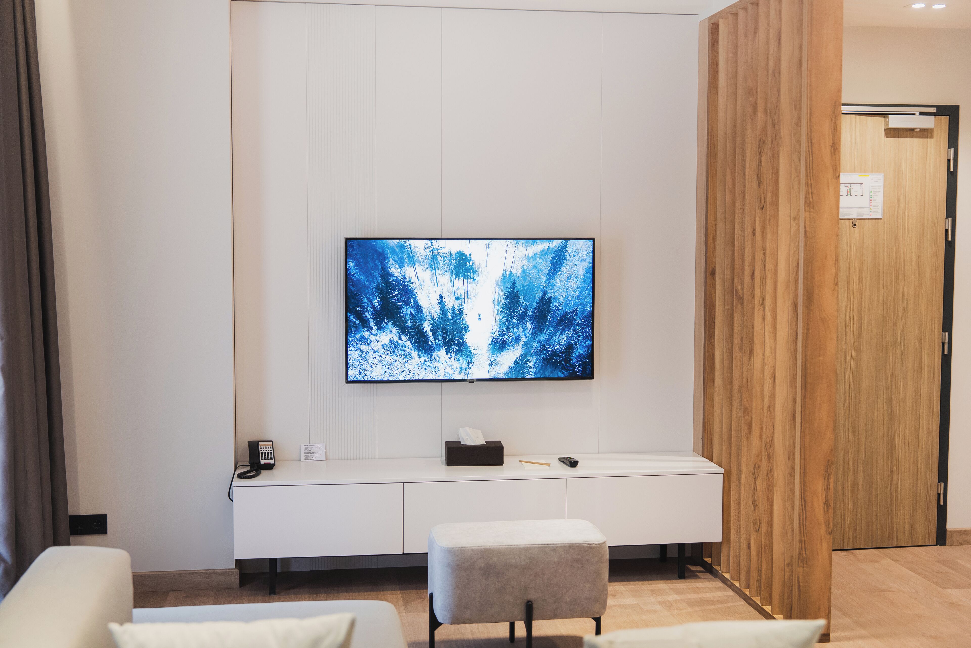 A modern hotel room with a large flat-screen TV mounted on the wall, a white console table with a telephone and tissues, and a grey ottoman.