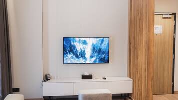 A modern hotel room with a large flat-screen TV mounted on the wall, a white console table with a telephone and tissues, and a grey ottoman.