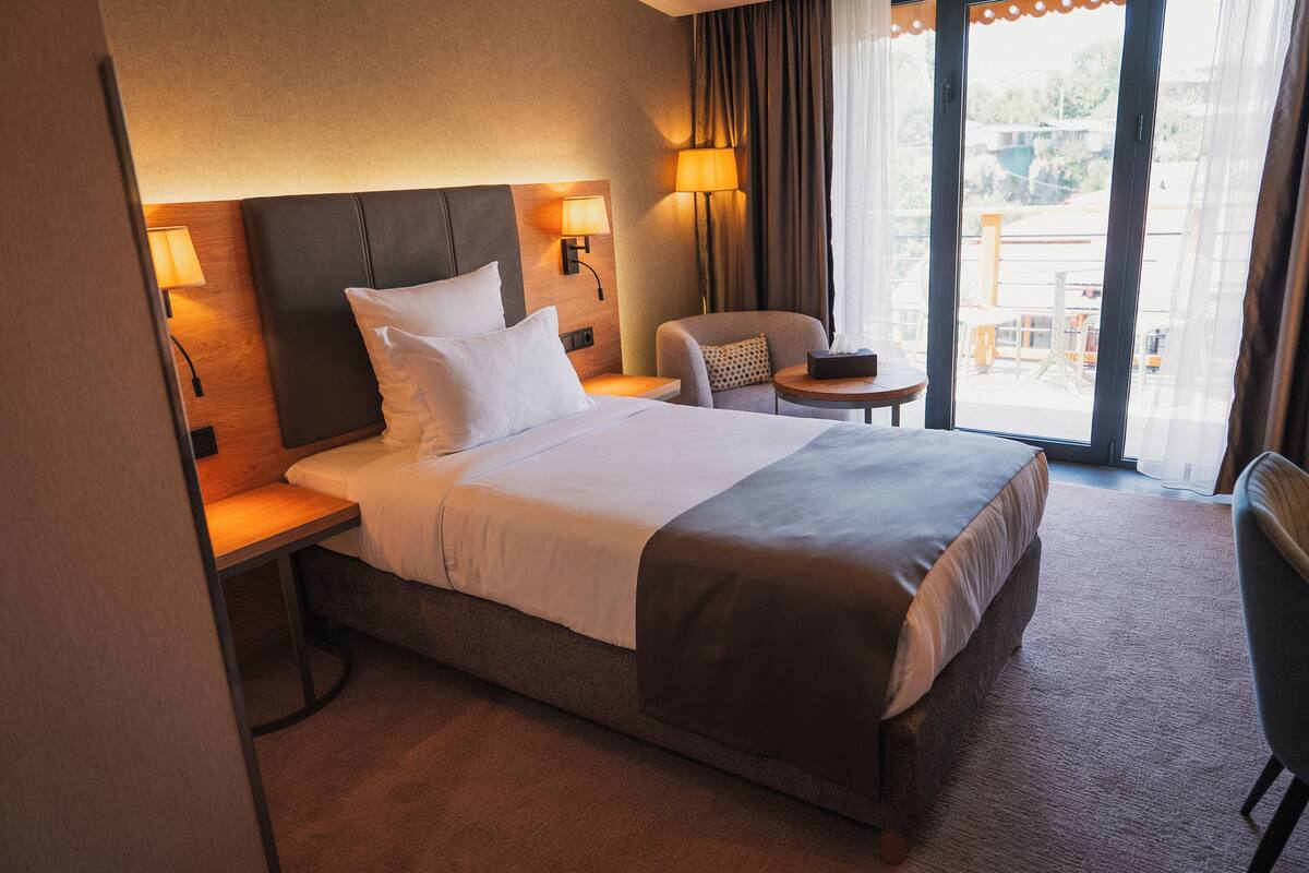 A hotel room with a large bed, a chair, a small table, and a view of the outdoors through a large window.