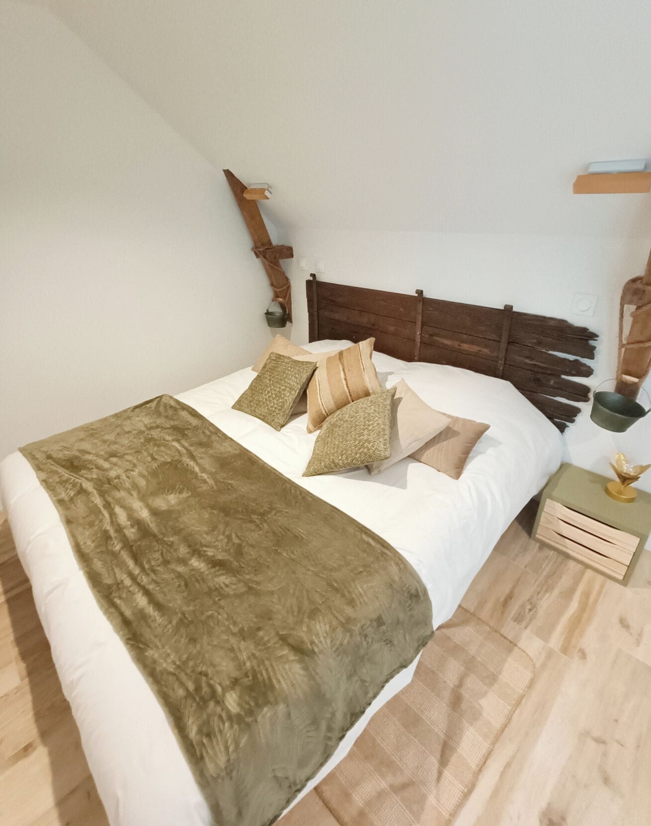 Ein Bett mit Holz-Kopfteil, mehreren Kissen, einer grünen Tagesdecke und einem Nachttisch aus Holz mit einer kleinen Lampe.