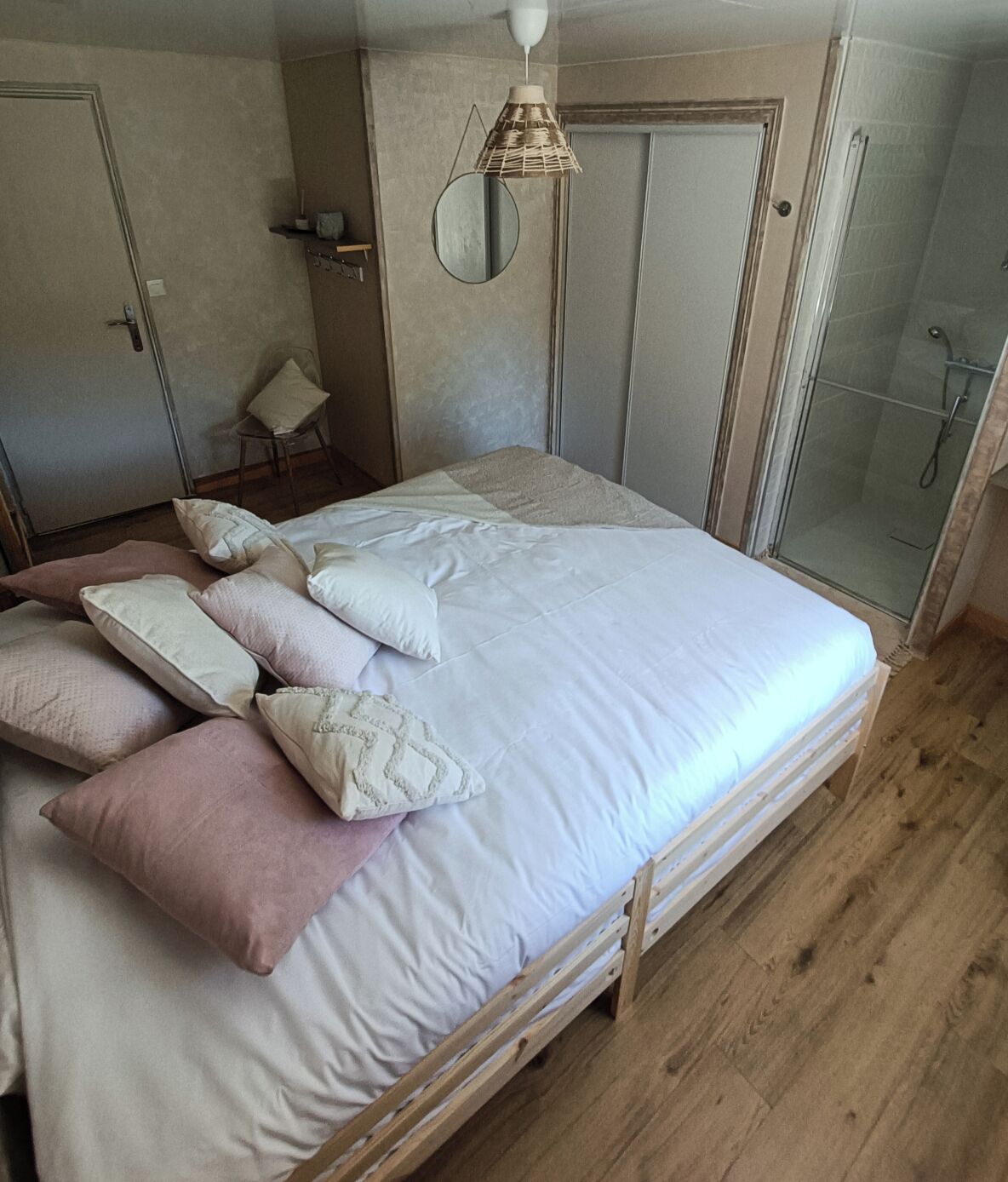 Een slaapkamer met een houten bed, witte en roze kussens, een hanglamp, een kleine stoel en een douchegedeelte.