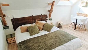 Un dormitorio con cabecera de madera, una cama grande con varias almohadas, una mesita de noche, una pequeña mesa redonda y una silla blanca. Hay un tragaluz sobre la cama.
